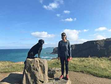 A walker smiles beside her dog, who sits atop a marker on the South West Coast Path walking holiday.