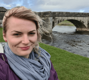 Gosia takes a selfie on the Three Dales Way with Burnsall Bridge, a stone bridge with multiple arches, in the background.