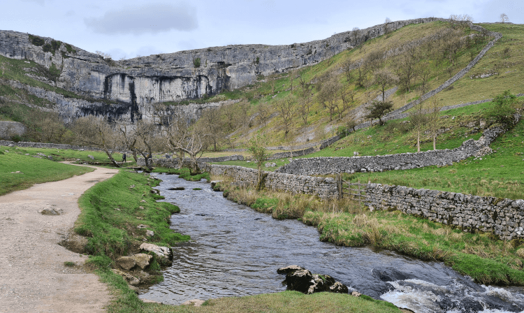 A view along the path leading to Malham Cove, itself a towering limestone cliff in the distance, with a dark stream bubbling away alongside.