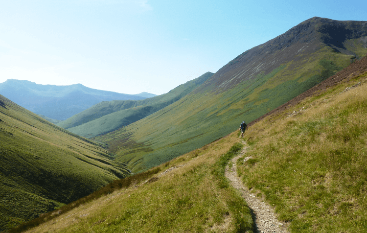 A walker follows a narrow path across a steep hillside between Buttermere and Newlands, with tall fells in the background.