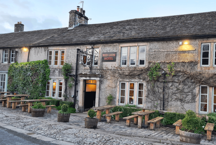 The Red Lion Pub on the Three Dales Way in Burnsall. It's a long stone pub with big three-paned windows and wooden benches outside, seen here in early evening.