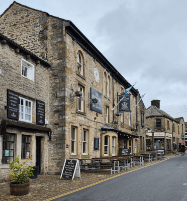 A view of The Devonshire pub in Grassington as viewed by Gosia on the Three Dales Way, made of honey-yellow stone with black accents and several picnic benches outside.