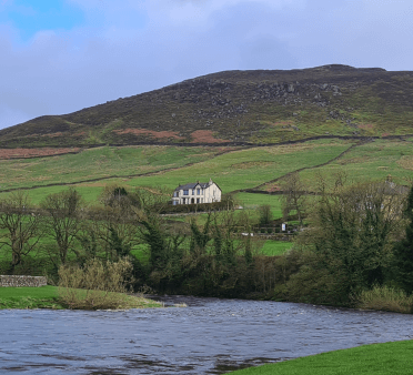 The views from Burnsall Bridge over the river and the green hills of Yorkshire beyond, with a white house sitting partway up the slope amongst fields split by low stone walls.