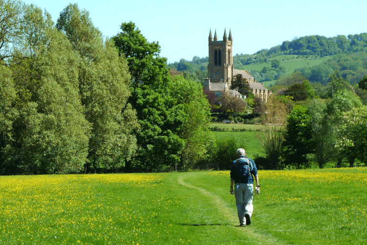 A walker crosses a green field on a beautiful spring day, with trees and a church in the distance as he approaches Broadway in the Cotswolds.