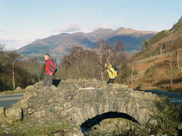 Walkers enjoying a waterside walk cross the sharply curved stone packhorse bridge of Ashness.