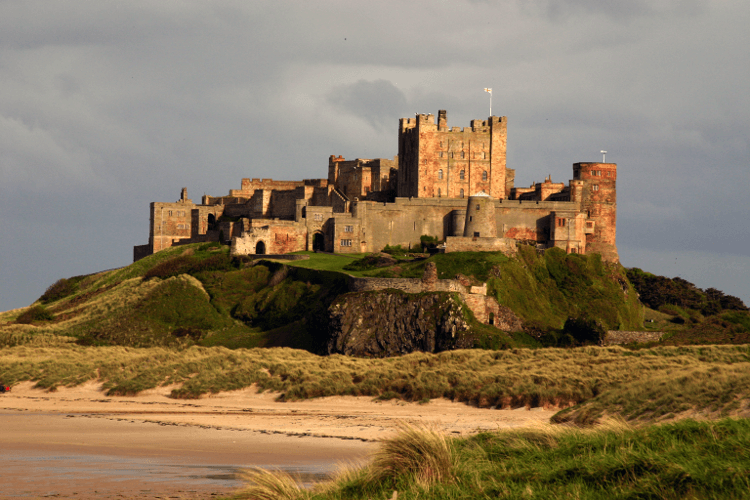 Bamburgh Castle sits on the UK coast, a large castle towering over the sand dunes below.