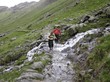 Two walkers, armed with waterproofs and walking sticks, cross a burn flowing across the path.