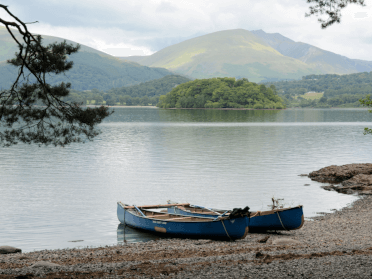 A wooden rowboat sits on the shore of Derwentwater, framed by pine trees.