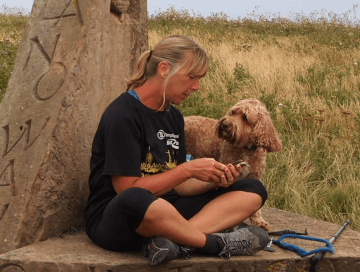 A walker sits on the Yorkshire Wolds Way monument on her dog-friendly walking holiday, checking the map as her poodle mix watches on.