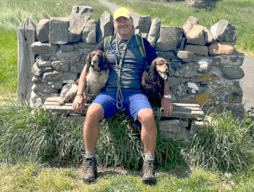 A walker sits on a stone bench with his arms around his spaniels, who sit on either side of him.