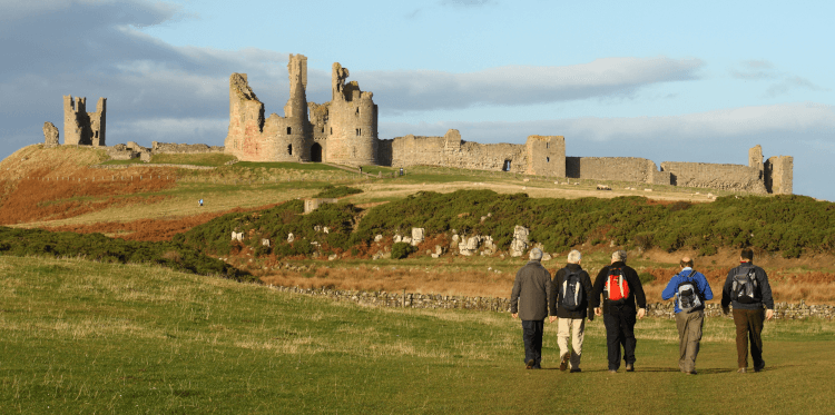 A group of walkers hike across grassy dunes towards Dunstanburgh Castle, a dramatic coastal ruin in Northumberland.