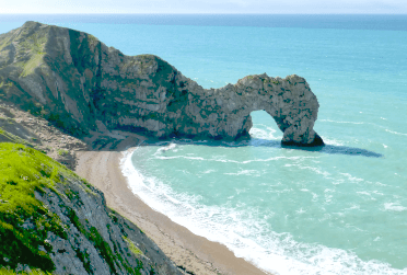 Durdle Door, an arch of rock standing out of the sea by the South West Coast Path, with a curve of beach in the foreground.