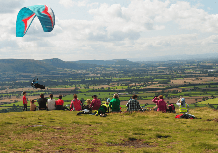 A large family walking group sit on the ground atop a grassy hill overlooking expansive views over the countryside on the Cleveland Way, as a hang glider passes above.