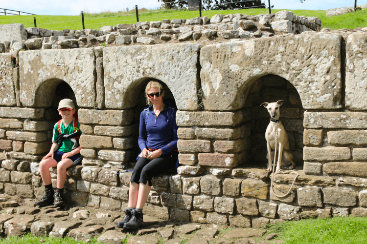 At a Roman fort on Hadrian's Wall Path, a child, adult and the family dog sit in stone alcoves on a bright sunny day, enjoying family fun on a long-distance hike.
