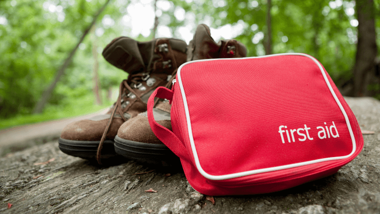 A first aid kit sits on a trail beside a good pair of hiking boots, ready to prevent blisters.