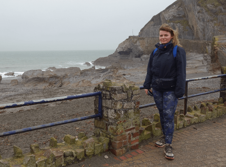 At the start of her walk from Ilfracombe to Woolacombe, Gosia stands in her walking gear near the sea on a blustery grey day.