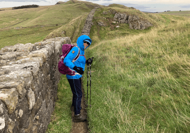 A walker in a blue coat walks alongside Hadrian's Wall, which stretches out ahead for miles.