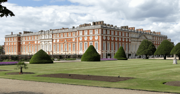 Hampton Court Palace from the Gardens by Maxwell Hamilton. Hampton Court Palace, an enormous building in red brick, stands above manicured gardens.