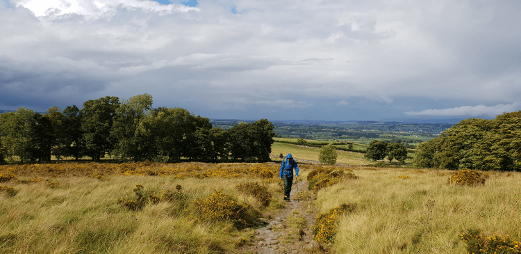 A lone hiker in blue waterproofs wanders along High Street Roman road, a grassy off-road path that was once a major thoroughfare.