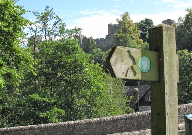 A wooden signpost for the Mortimer Trail with the silhouette of a castle in the background through the trees