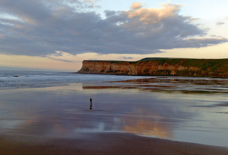 A walker takes time to ground themselves with the sounds and sights of the surf on the Yorkshire Coast, with a projecting headland in the background reflected in the still water left by the receding tide.