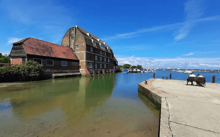 A view across the still blue water of a harbour to a red brick industrial building opposite.