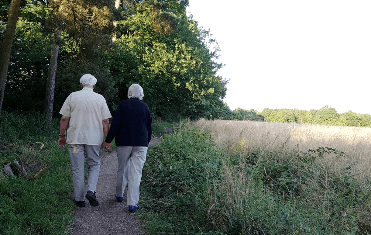 Elderly parents walk along a footpath beside a wood.