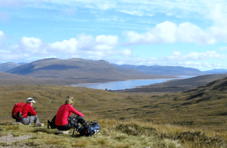 Two walkers sit on the ground on a brisk winter's day to enjoy a break and a snack. The landscape is very open, all the way out to a broad lake and distant hills.