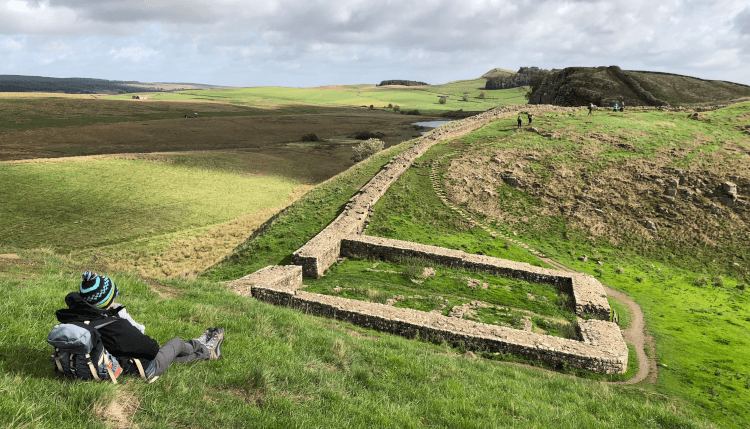 A solo hiker sits with their pack on Hadrian's Wall Path, overlooking the trail and Roman ruins.