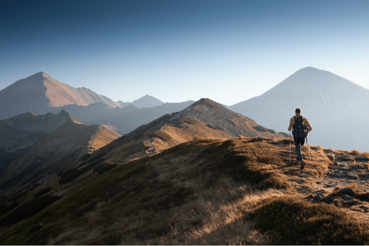 A man with walking poles follows a ridgetop path through the mountains.