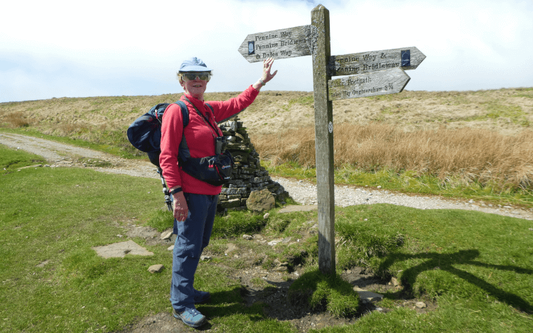 A walker stands on a grassy trail in the countryside holding a marked-out map.