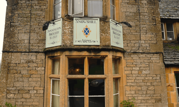 Snowshill Arms by Bex Walton. A close-up of the Snowshill Arms, a honey-stone Cotswold pub with a hand-painted sign hoisted on the wall between two bay windows.