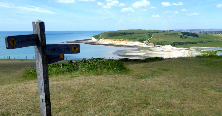 A fingerpost for the South Downs Way points along the English coastline, following a narrow track through grassy fields with the cliff edge beyond and the white cliffs in the distance.