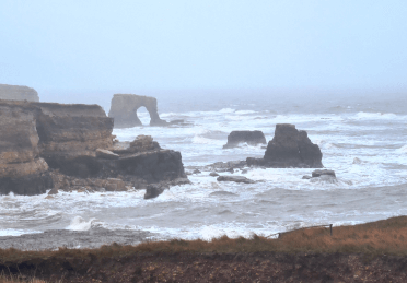 A view down to a stone arch and headland through the storm haze shrouding the South Northumberland Coast Path.