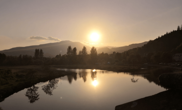 Sunset over the trail. The sun sinks beneath the hills, with trees silhouetted and the view mirrored in a still lake at the bottom of the photo.