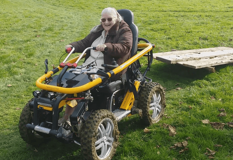 An older woman sits in a Tramper all-terrain hopper, a mobility scooter with long travel suspension and knobbly tyres.
