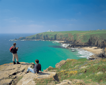 Two walkers relax on the South West Coast Path overlooking a wide azure bay and Lizard Point.
