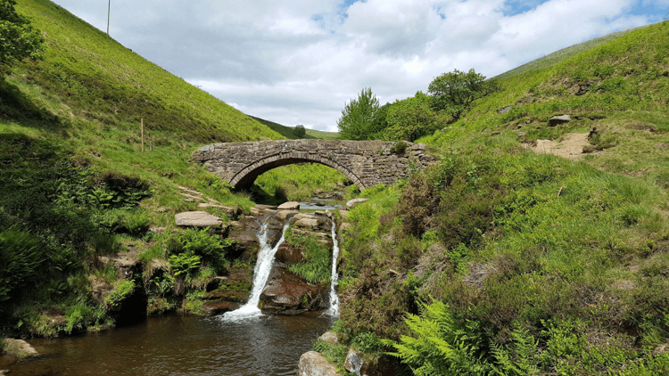 Dane Valley Way at Three Shires Head by Douglal. A waterfall runs beneath an arched stone bridge in lush green scenery.