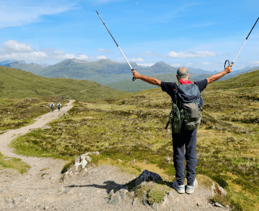 At the top of Devil's Staircase, a challenging climb in remote Scotland, a walker stands with poles raised in a victory pose.