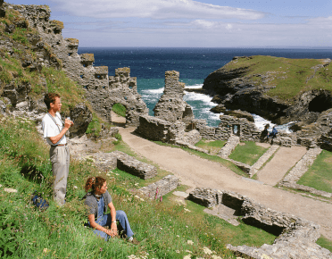 Two walkers sit amongst the grass and stone of the ruins of Tintagel Castle.