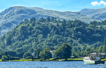 A boat crosses the water beneath fell peaks in the Lake District, home to incredible waterside walks.